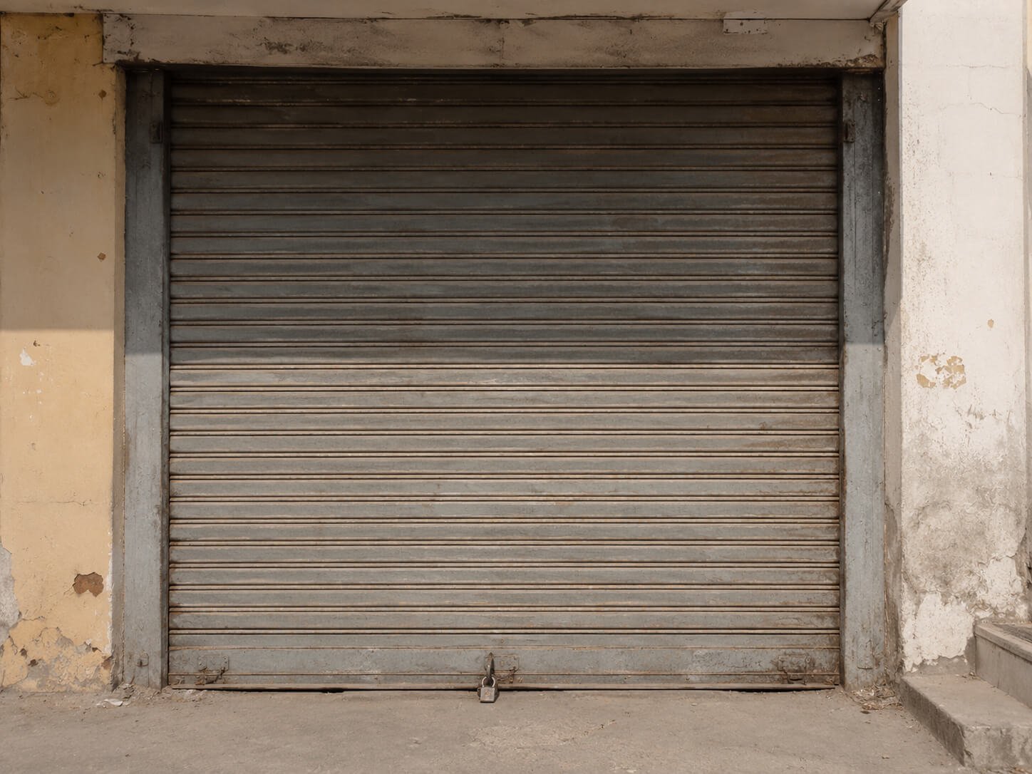 The closed metal shutter of a small commercial unit, padlocked, in flat daylight