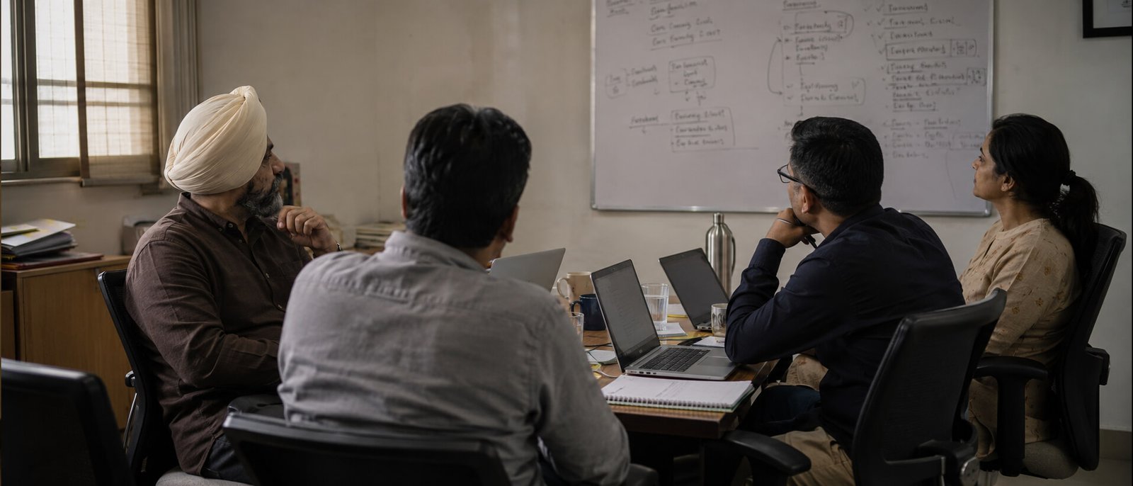 A working meeting room in a mid-sized company, four people at a table with a whiteboard of messy handwriting behind them