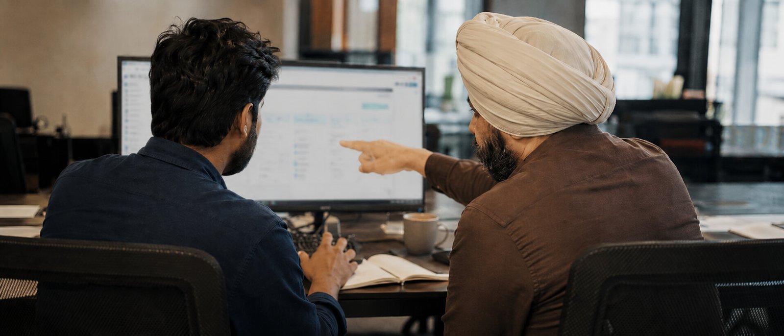 Two colleagues working shoulder to shoulder at one monitor, one typing and one pointing at the screen