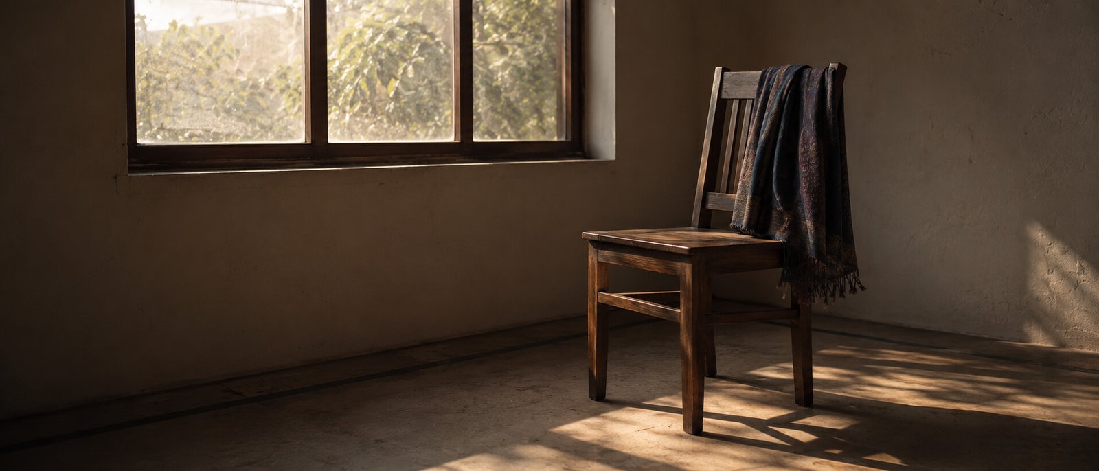 An empty wooden chair beside a window in early morning light, a folded shawl over the back